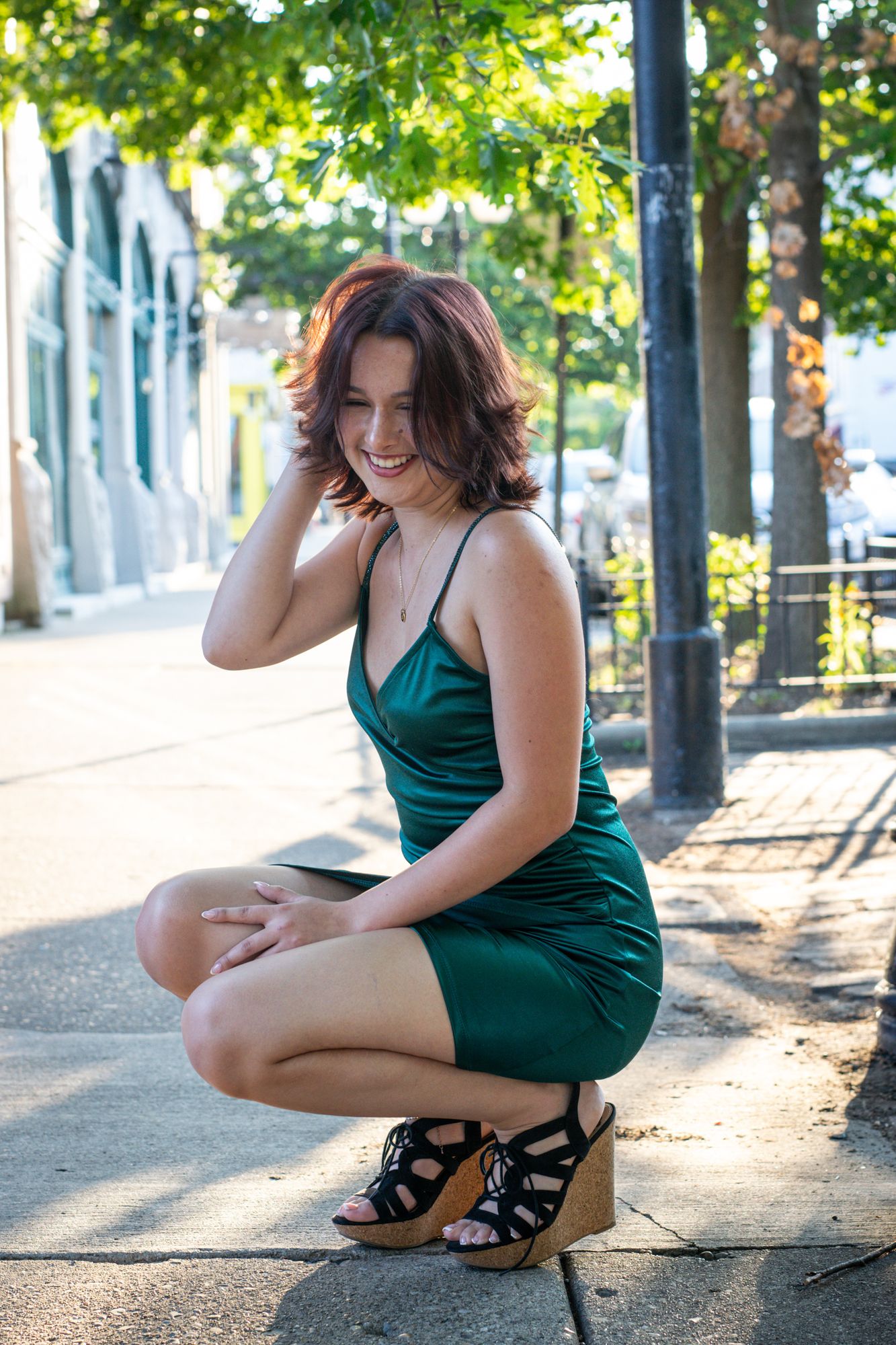 A senior crouched on a tree-lined sidewalk, laughing, backlit by late afternoon light