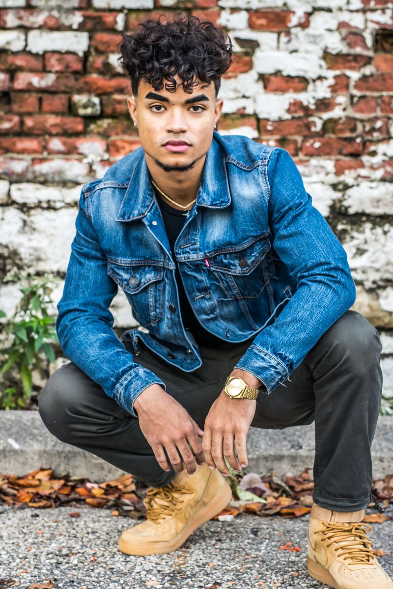 A senior in a denim jacket crouched against a weathered painted brick wall, autumn leaves at his feet